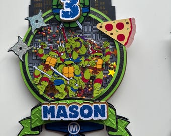May include: A colorful cake topper featuring the Teenage Mutant Ninja Turtles. The design includes a green number 3, a pizza slice, and the name "MASON". The background has a cityscape and ninja stars.