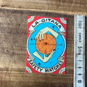 Op de afbeelding: Een vintage etiket voor een luciferdoos met een blauwe en rode achtergrond. Het etiket toont een gestileerde illustratie van twee handen die een bol met een oog in het midden vasthouden. De tekst "LA GITANA SAFETY MATCHES" is in zwarte letters op het etiket gedrukt.