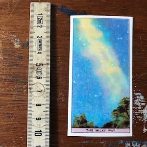 Puede incluir: Una ilustración vintage de la Vía Láctea, una banda de estrellas y luz contra un cielo nocturno azul. La imagen está impresa en una tarjeta blanca con el texto "THE MILKY WAY" en la parte inferior.