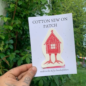 Peut inclure: Un écusson à coudre en coton représentant une maison rouge sur pattes. Le patch est présenté sur une carte blanche avec l'inscription "COTTON SEW ON PATCH". La carte porte également la mention "made in the uk by lotusleafaltars".