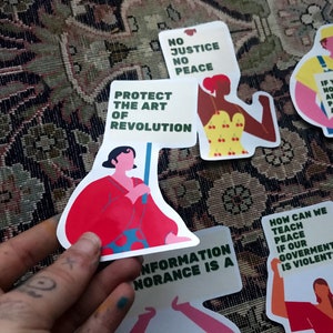 Pu&ograve; includere: Un set di adesivi colorati con messaggi femministi e politici. Gli adesivi presentano illustrazioni di donne che tengono cartelli con slogan come "Proteggere l'arte della rivoluzione", "Nessuna giustizia, nessuna pace" e "Come possiamo insegnare la pace se il nostro governo &egrave; violento?"
