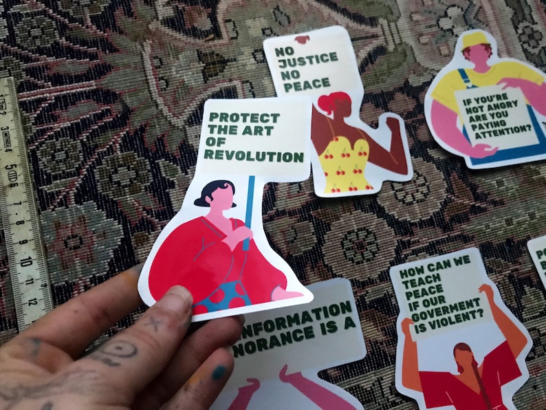 Pu&ograve; includere: Un set di adesivi colorati con messaggi femministi e politici. Gli adesivi presentano illustrazioni di donne che tengono cartelli con slogan come "Proteggere l'arte della rivoluzione", "Nessuna giustizia, nessuna pace" e "Come possiamo insegnare la pace se il nostro governo &egrave; violento?"