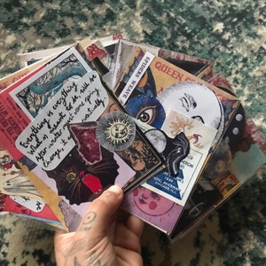 May include: A stack of colorful stickers with various designs, including a black cat, a moon, a spider web, and a quote that reads "Everything is everything. What is meant to be, will be. After winter must come spring. Change, it eventually."