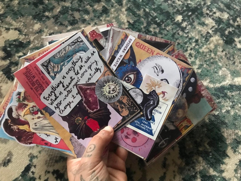 May include: A stack of colorful stickers with various designs, including a black cat, a moon, a spider web, and a quote that reads "Everything is everything. What is meant to be, will be. After winter must come spring. Change, it eventually."