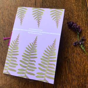 Op de afbeelding: Een lavendelkleurig boekje met een varenspatroon en de titel "A Calendar for Wildcrafting and Foraging in the Northern Hemisphere" door "Botanical Folk"