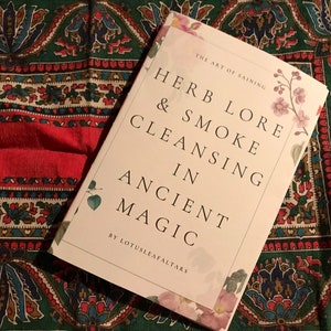 Puede incluir: Un libro titulado "Herb Lore & Smoke Cleansing in Ancient Magic" de Lotusleafaltars. El libro tiene una cubierta blanca con detalles florales y está sobre una tela estampada verde y roja.