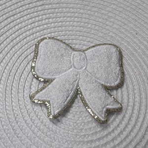 Peut inclure: Un nœud en tissu blanc avec un contour doré pailleté. Le nœud est posé sur une surface blanche texturée. Le design comprend un nœud central et des extrémités bouclées, créant un accent décoratif.