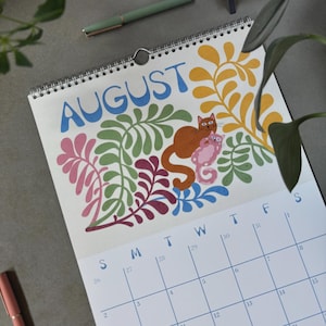 May include: A spiral-bound calendar open to August, featuring a colorful illustration of a cat and a seahorse amidst floral designs. The word "AUGUST" is in blue script. The calendar grid is visible with blue numbers and days.