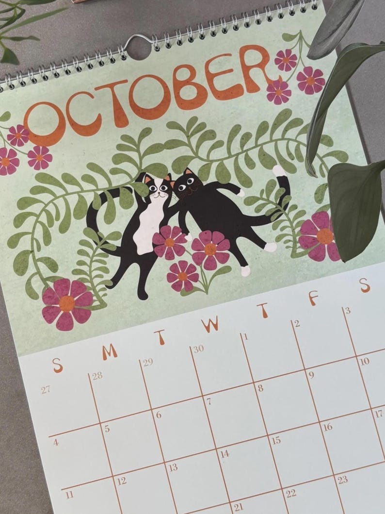 May include: A spiral-bound calendar for October featuring a light green background with orange text. The calendar has illustrations of two black cats with white markings, surrounded by pink flowers and green foliage. The calendar grid is visible.