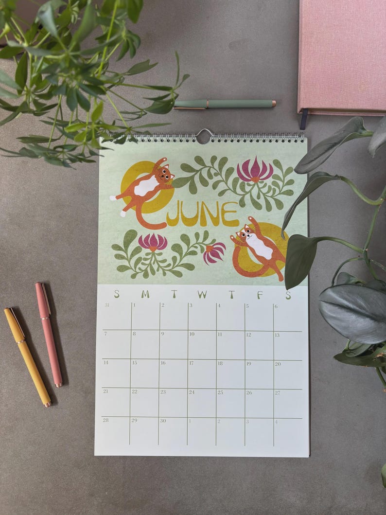 May include: A wall calendar for June featuring illustrations of orange and white cats on yellow circles, surrounded by floral designs. The word "JUNE" is in a gold font. The calendar is displayed with pens and a notebook on a gray surface.