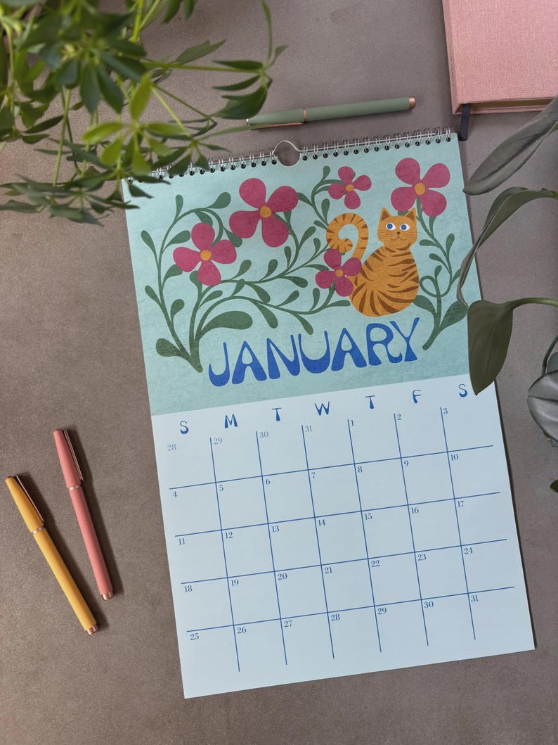 May include: A spiral-bound calendar for January featuring a light blue background with pink flowers and a cartoon orange tabby cat. The word "JANUARY" is in blue. Two pens are visible, one yellow and one pink.