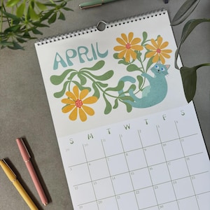 May include: A spiral-bound calendar for April featuring a teal cat among yellow flowers and green leaves. The word "APRIL" is in teal script. Three pens in yellow, pink, and green are on the gray surface.