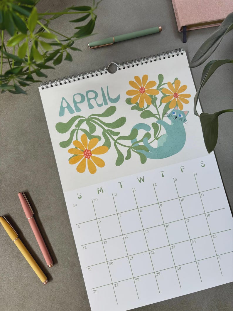 May include: A spiral-bound calendar for April featuring a teal cat among yellow flowers and green leaves. The word "APRIL" is in teal script. Three pens in yellow, pink, and green are on the gray surface.