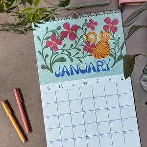 May include: A wall calendar for January featuring a light blue background with a cartoon orange tabby cat and pink flowers. The word "JANUARY" is in blue. Three pens in yellow and pink are on the left.