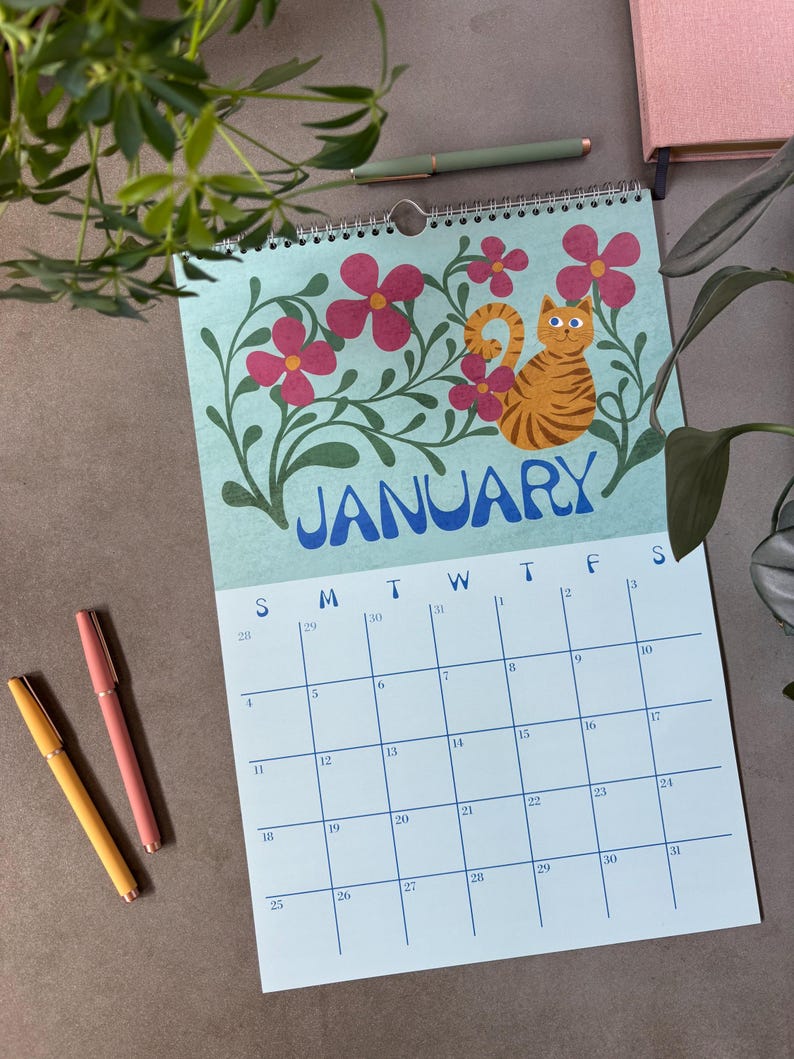 May include: A wall calendar for January featuring a light blue background with a cartoon orange tabby cat and pink flowers. The word "JANUARY" is in blue. Three pens in yellow and pink are on the left.