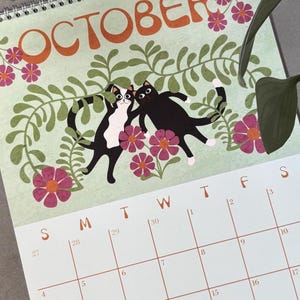 May include: A spiral-bound calendar page for October featuring a light green background with orange text. The design includes two cartoon cats, flowers, and foliage. The calendar grid is visible below the illustration.