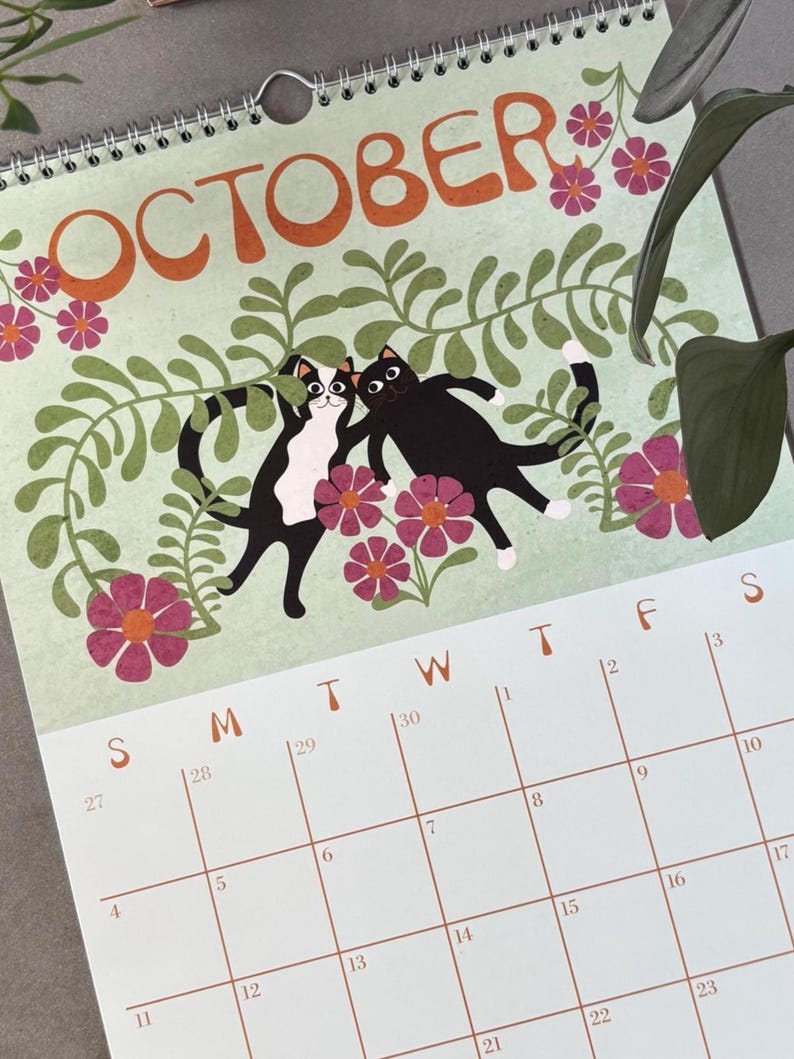May include: A spiral-bound calendar page for October featuring a light green background with orange text. The design includes two cartoon cats, flowers, and foliage. The calendar grid is visible below the illustration.