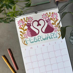 May include: A spiral-bound calendar open to February, featuring two stylized pink cats forming a heart shape. The word "FEBRUARY" is in teal script. Pink and yellow pens and greenery are in the background.