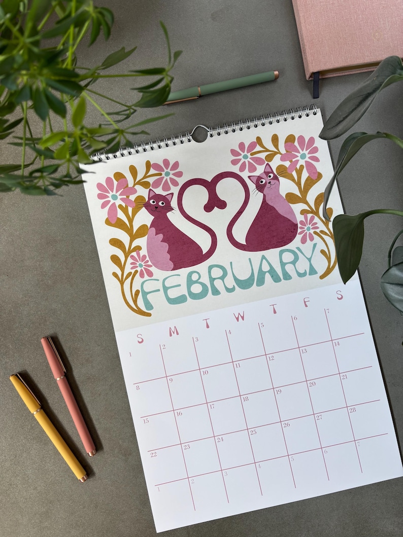 May include: A spiral-bound calendar open to February, featuring two stylized pink cats forming a heart shape. The word "FEBRUARY" is in teal script. Pink and yellow pens and greenery are in the background.