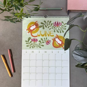 May include: A wall calendar for June with a light green background, featuring illustrations of orange and white cats. The word "JUNE" is in gold lettering. The calendar is surrounded by floral designs and a grid for the days of the month.