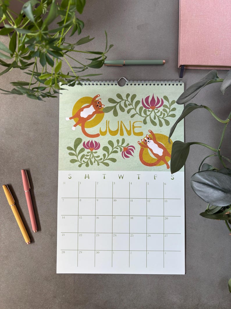 May include: A wall calendar for June with a light green background, featuring illustrations of orange and white cats. The word "JUNE" is in gold lettering. The calendar is surrounded by floral designs and a grid for the days of the month.