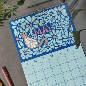 May include: A spiral-bound calendar featuring a blue background with white and pink flowers. The word "MAY" is in pink. A gray cat with a white belly is lounging on the top portion. The calendar grid is light blue.