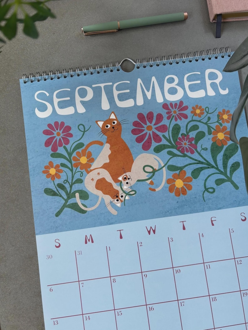 May include: A spiral-bound calendar for September with a light blue background. The calendar features a whimsical illustration of orange and white cats among colorful flowers. The word "SEPTEMBER" is at the top.