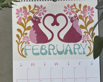 May include: A wall calendar for the month of February featuring a graphic illustration of two pink and purple cats with tails forming a heart shape, surrounded by gold and pink flowers. The calendar grid displays the days from 1 to 28.