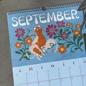 May include: A spiral-bound calendar for September with a light blue background. The calendar features an illustration of orange and white cats among flowers. The word "SEPTEMBER" is at the top. A green pen rests on the surface.