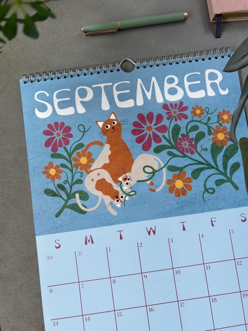 May include: A spiral-bound calendar for September with a light blue background. The calendar features an illustration of orange and white cats among flowers. The word "SEPTEMBER" is at the top. A green pen rests on the surface.