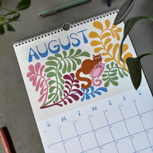 May include: A spiral-bound calendar open to August, featuring a colorful illustration of a cat and a kitten amidst floral designs. The word "AUGUST" is in blue, with a calendar grid below. A green pen and a pink notebook are visible.
