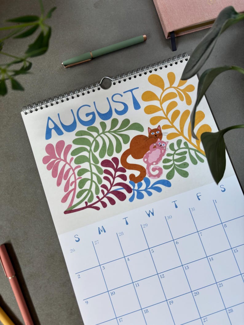 May include: A spiral-bound calendar open to August, featuring a colorful illustration of a cat and a kitten amidst floral designs. The word "AUGUST" is in blue, with a calendar grid below. A green pen and a pink notebook are visible.
