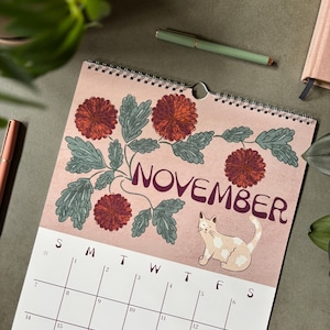 May include: A wall calendar for November featuring a pink background with dark red chrysanthemum flowers, green leaves, and a light-colored cat. The calendar grid displays the days of the week and dates in black ink on a white background with a spiral binding.