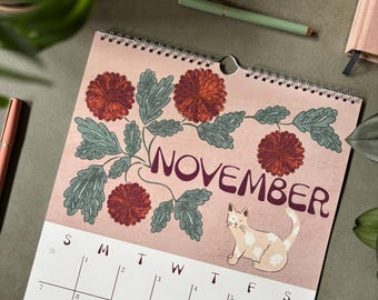May include: A wall calendar for November featuring a pink background with dark red chrysanthemum flowers, green leaves, and a light-colored cat. The calendar grid displays the days of the week and dates in black ink on a white background with a spiral binding.