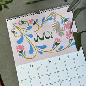 May include: A spiral-bound calendar for July, featuring a pink background with floral and cat illustrations. The word "JULY" is in green, and the calendar grid is visible below. A green pen and notebook are in the background.