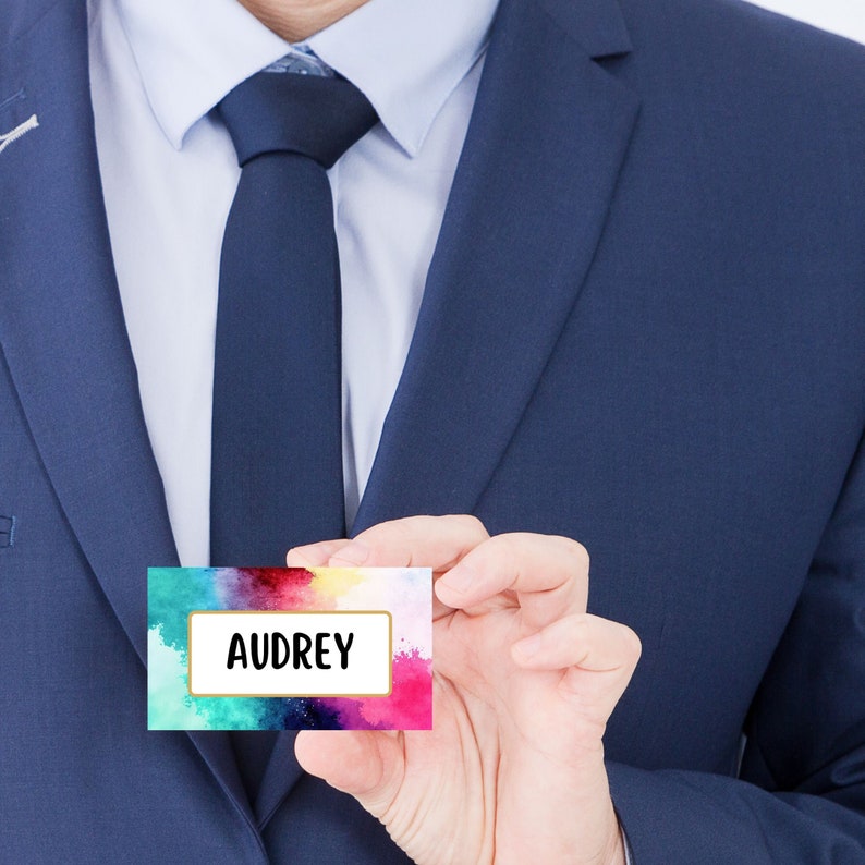 May include: A person in a blue suit holds a colorful watercolor name tag with the name "AUDREY" printed in gold.