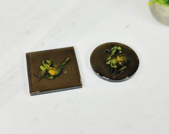 Puede incluir: Dos posavasos marrones con un diseño de flores prensadas. Un posavasos es cuadrado y el otro es redondo. Las flores son verdes y amarillas.
