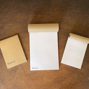 K&ouml;nnte beinhalten: Drei braune Papiernotizb&uuml;cher mit liniertem wei&szlig;em Papier. Das obere Notizbuch tr&auml;gt den Namen "Karen" auf der Vorderseite. Das untere linke Notizbuch tr&auml;gt den Namen "Karen" auf der Vorderseite. Das untere rechte Notizbuch ist leer.