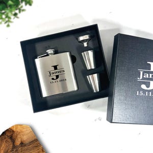 Puede incluir: Una petaca de plata con una inicial "J" grabada en negro y el nombre "James" con la fecha "15.11.2024". La petaca está en una caja negra con dos vasos de chupito de plata y un embudo.