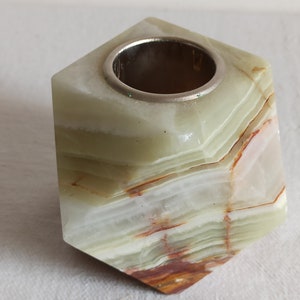 Puede incluir: Un portavelas de &oacute;nix verde, blanco y marr&oacute;n con un anillo de metal plateado en la parte superior. El portavelas tiene forma de cubo con bordes redondeados.