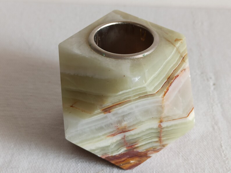 Puede incluir: Un portavelas de &oacute;nix verde, blanco y marr&oacute;n con un anillo de metal plateado en la parte superior. El portavelas tiene forma de cubo con bordes redondeados.