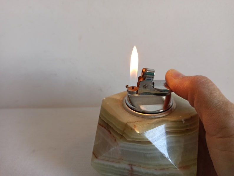 Puede incluir: Un encendedor de mesa de m&aacute;rmol verde y blanco con un encendedor plateado en la parte superior. El encendedor est&aacute; encendido y la llama es visible.