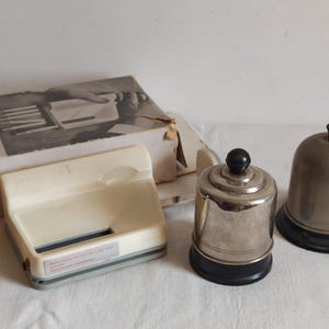 May include: An assortment of vintage items, including a cream-colored paper cutter, a silver-toned bell with a black knob, and a matching bell. A box with the word "PRIVILEGE" is also visible.