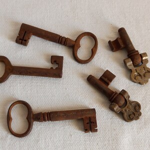 Peut inclure: Cinq clés anciennes avec une finition brun rouillé sont disposées sur une surface blanche. Les clés varient en taille et en design, avec des têtes de clés ornées et simples. Les clés sont probablement vintage ou décoratives.