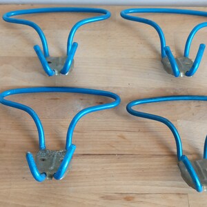 May include: Four vintage blue metal wall hooks. Each hook has a curved top and two downward-facing prongs, attached to a small, rectangular metal base. The hooks are arranged on a light-colored wooden surface.