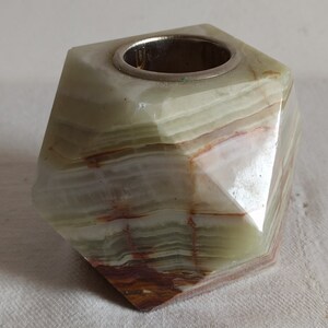 Puede incluir: Un portavelas de &oacute;nix verde, blanco y marr&oacute;n con un inserto de metal plateado. El portavelas tiene forma de prisma geom&eacute;trico facetado.