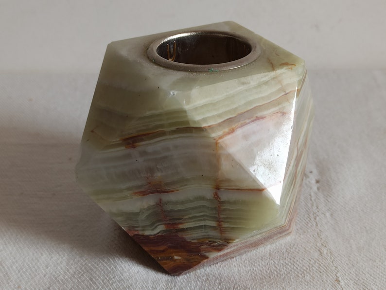 Puede incluir: Un portavelas de &oacute;nix verde, blanco y marr&oacute;n con un inserto de metal plateado. El portavelas tiene forma de prisma geom&eacute;trico facetado.