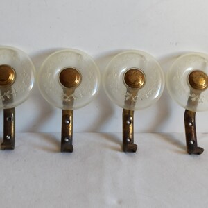May include: Four vintage wall hooks. Each hook features a clear, circular glass knob with a floral design, a brass-colored center, and a dark metal bracket. The hooks are arranged in a row, showcasing their design and construction.