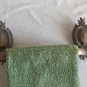 May include: An antique towel rail with a green towel. The rail has ornate, oval-shaped bronze-coloured mounts with floral accents. The towel is a solid, textured green.