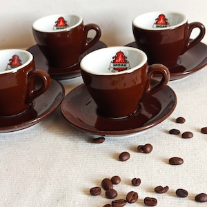 May include: Four espresso cups and saucers in a rich brown color. Each cup has a white interior with a logo and sits on a matching saucer. Coffee beans are scattered around the cups, suggesting a coffee-related theme.